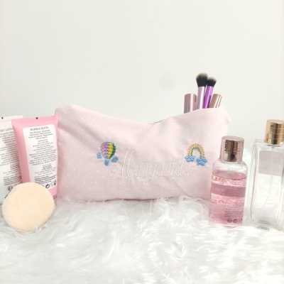 Necessaire rosa clara bordada com balão e arco-íris, pincéis de maquilhagem e frascos transparentes sobre superfície branca felpuda