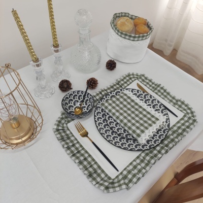 Mesa posta com prato, tigela, talheres, guardanapo xadrez, castiçais dourados com velas, cesta de pão e toalha branca.