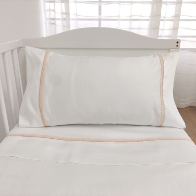 Fronha e lençol branco com detalhe cor de rosa claro numa cama branca junto a uma janela com cortinas brancas