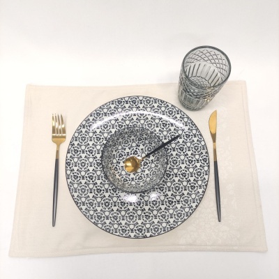 Conjunto de louça com prato e taça branco com padrão azul, talheres pretos e dourados, copo transparente com padrão, em individual branco.