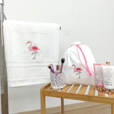 Toalha branca com flamingo bordado, saco de tecido branco com flamingo, pincéis de maquilhagem e cremes sobre banco de madeira.