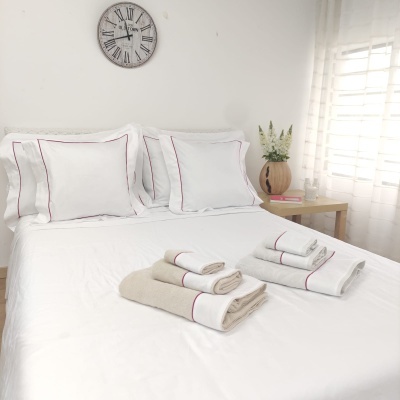 Cama com lençóis, fronhas e toalhas brancos e bege com costura vermelha, relógio de parede e mesa com vaso e livros