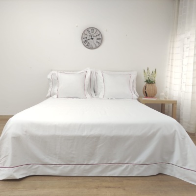 Cama de casal com roupa de cama branca e linha vermelha, relógio na parede, mesa de cabeceira com vaso e planta, cortina branca