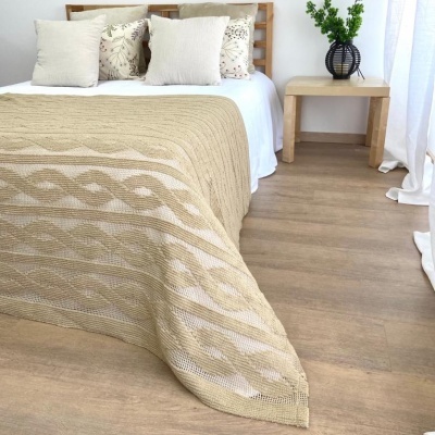 Cobertor bege com padrão trançado em relevo sobre cama branca com almofadas bege e creme
