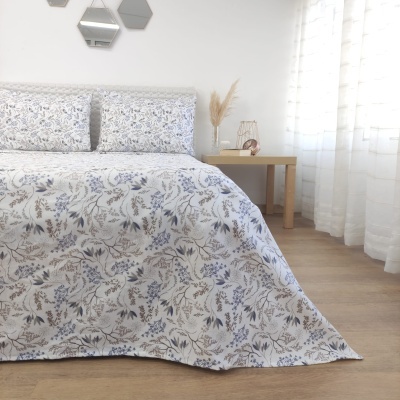 Cama com conjunto de lençol e fronhas florais cinza, azul e bege num quarto com piso de madeira e cortinas brancas