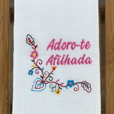 Toalha branca bordada com texto 'Adoro-te Afilhada' e flores coloridas sobre madeira