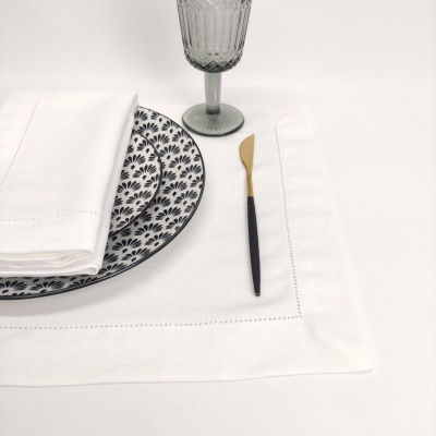 Conjunto de jantar com prato decorado, guardanapo branco, talher preto e dourado, copo acinzentado e toalha branca.