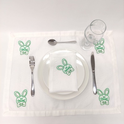 Jogo americano branco com coelhos bordados verdes, pratos brancos, talheres metálicos e copo de vidro
