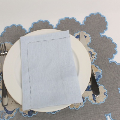 Mesa posta com prato branco, guardanapo azul claro, talheres metálicos e individual cinza com bordas florais azuis.