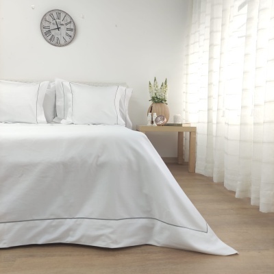 Cama com colcha e fronhas brancas, relógio de parede, cortinas brancas translúcidas, mesa de madeira com vaso de flores