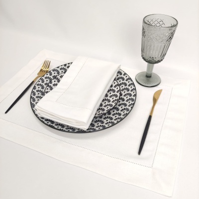 Conjunto de mesa com prato cerâmico preto e branco, guardanapo e toalhete brancos, talheres preto e dourado, e copo cinzento transparente