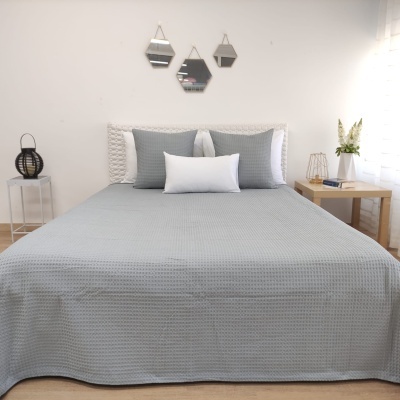 Cama de casal com colcha cinzenta texturada e almofadas, cabeceira trançada branca, mesa de cabeceira com vaso de flores, três espelhos hexagonais na parede.