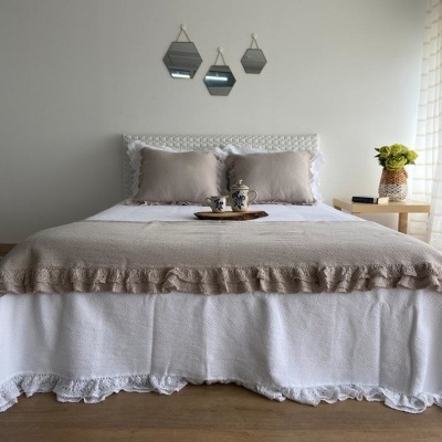 Quarto com cama de casal decorada com colcha branca e manta bege, almofadas, espelhos hexagonais e mesa com vaso