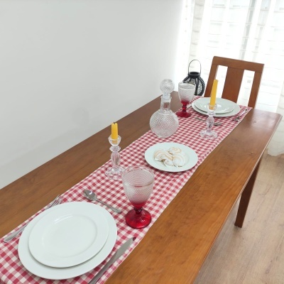 Mesa de jantar com toalha xadrez vermelha e branca, pratos brancos, taças vermelhas, castiçais com velas amarelas e jarro de cristal.