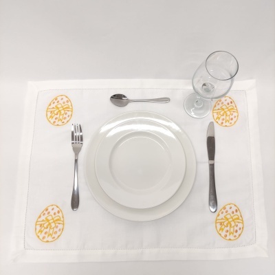 Mesa posta com pratos brancos, talheres metálicos, copo de vidro e toalha branca com desenhos de ovos amarelos