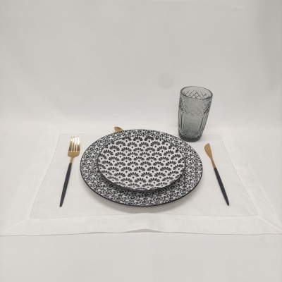 Conjunto de jantar com pratos preto e branco, talheres pretos com dourado e copo cinza sobre jogo americano branco.