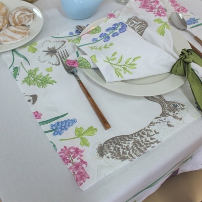Toalha e guardanapo de tecido branco com estampas coloridas de flores, borboleta e lebre, com talheres e prato branco