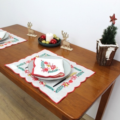 Jogo americano e guardanapo natalícios bordados em tecido branco com decoração verde e vermelha sobre mesa de madeira com detalhes de Natal