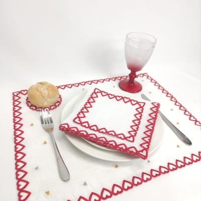 Conjunto de tecido branco com bordado vermelho, prato branco, talheres metálicos, copo vermelho e pão sobre suporte