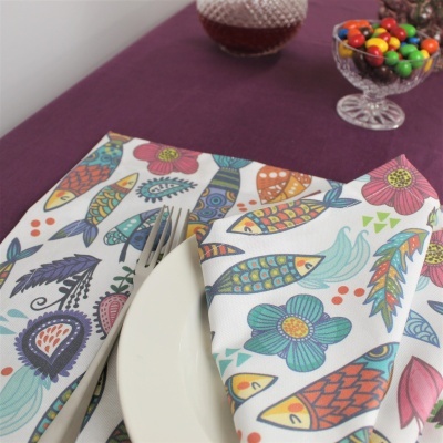 Guardanapos de tecido com padrão colorido de peixes e flores sobre mesa lilás com prato branco e taças