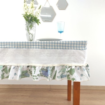 Toalha de mesa azul clara com babado floral e faixa branca em mesa de madeira com copo azul e vaso de flores