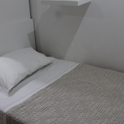 Cama individual com roupa de cama branca e manta cinza em quarto simples