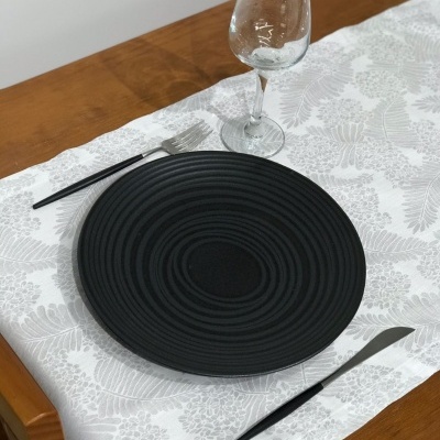 Mesa posta com prato preto, talheres pretos e copo de vidro em toalha branca com padrão floral.