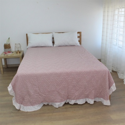 Cama com colcha rosa com babado branco e almofadas brancas junto a mesa de cabeceira e cortina branca