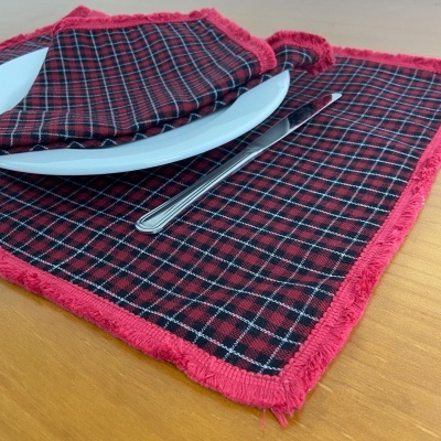 Guardanapo e toalhete xadrez vermelho com bordas vermelhas e franja sobre mesa de madeira com prato branco e faca.