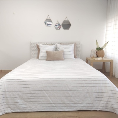 Cama com roupa de cama branca às riscas cinza, cabeceira almofadada cinza, três espelhos geométricos na parede e mesa de madeira com decoração