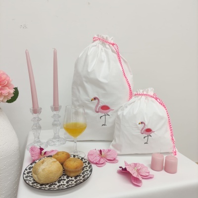 Dois sacos de tecido branco com estampas de flamingo rosa e cordão rosa, pão e muffin em prato, taça com bebida amarela e velas rosas em superfície branca.