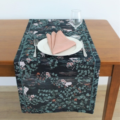 Passadeira de mesa com padrão de folhas e animais sobre mesa de madeira com louça branca e copo