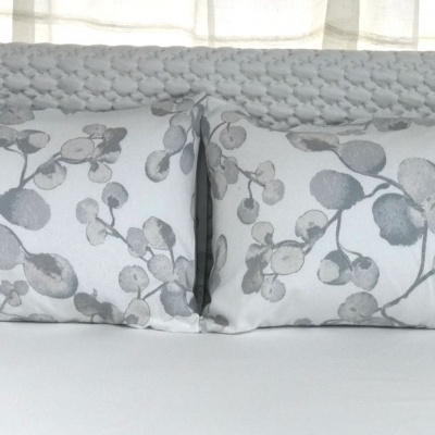 Cabeceira cinzenta trançada, almofadas com padrão folhas cinzentas, cama branca com lençol branco.