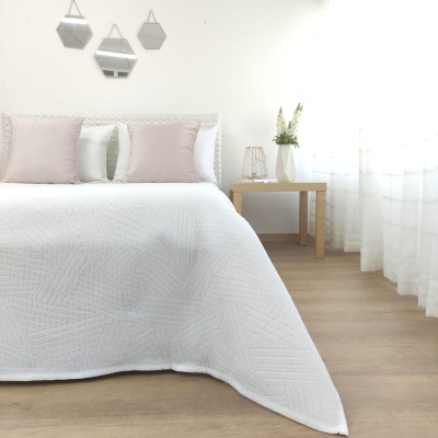 Cama com colcha branca acolchoada e almofadas rosa e brancas, mesa de madeira com decoração, espelhos hexagonais na parede, chão em madeira clara