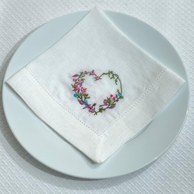 Guardanapo branco com bordado de coração floral num prato branco sobre toalha branca texturizada