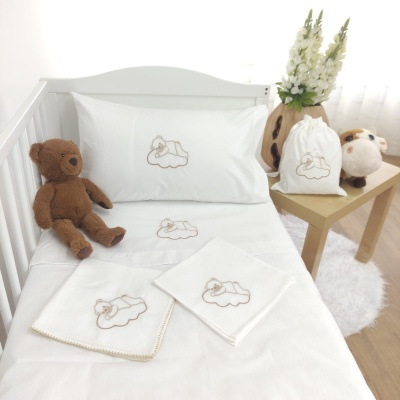 Roupa de cama branca para berço com bordado de ursinho, ursinho de peluche castanho, saco branco, peluche castanho e branco na mesa, jarro com flores brancas.