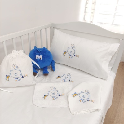Conjunto de roupa de cama e acessórios para bebé com ursinho azul bordado num berço branco.