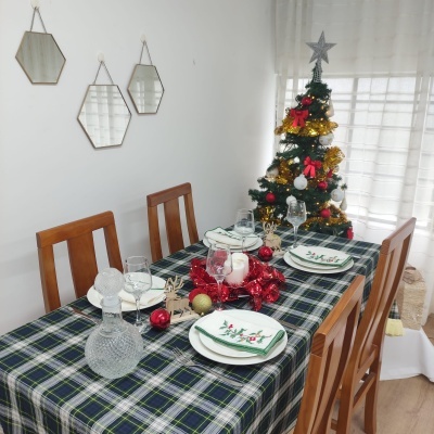 Mesa de jantar decorada para Natal com toalha tartan, pratos com motivos natalícios, taças e árvore de Natal ao fundo