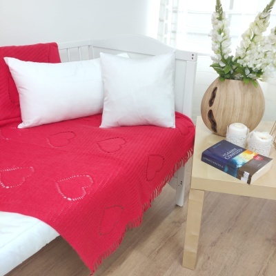 Colcha vermelha com corações bordados sobre cama branca com almofadas brancas e mesa de madeira ao lado