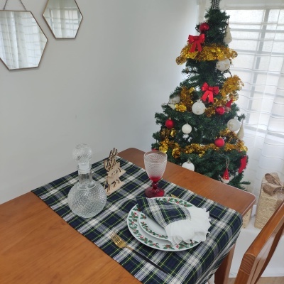 Mesa posta com toalha xadrez, prato decorado, copo e garrafa de vidro, próximo a uma árvore de Natal decorada