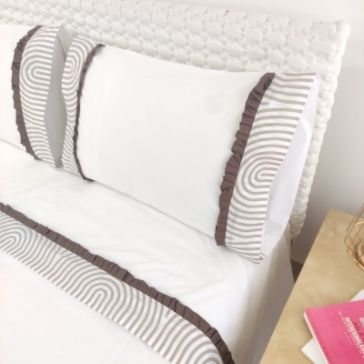 Cama com almofadas brancas e detalhes cinzentos e castanhos, cabeceira acolchoada branca e mesa de cabeceira clara com objetos