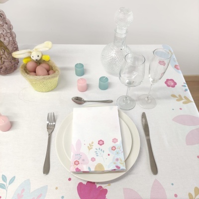 Mesa posta com prato, talheres, guardanapo floral, copos e decantador de vidro, velas coloridas e cesta com ovos e coelho.