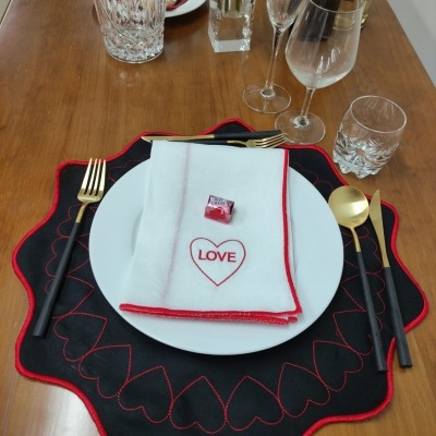 Conjunto de jantar com toalha preta bordada, prato branco, guardanapo com LOVE, talheres e copos sobre mesa de madeira