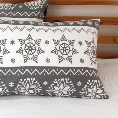 Travesseiro decorativo cinzento e branco com padrão de flocos de neve, em cama com cabeceira de madeira e colcha branca.