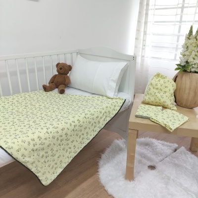 Cobertor amarelo claro com padrão floral numa cama branca e acessórios correspondentes sobre mesa em quarto iluminado