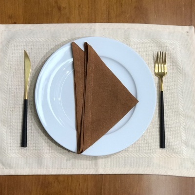 Conjunto de jantar com prato branco e talheres dourados e pretos sobre individual bege e mesa de madeira