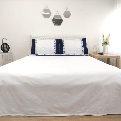 Cama de casal com roupa branca e detalhes azuis, três espelhos geométricos na parede, mesa de cabeceira com flores e decoração