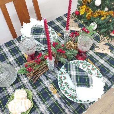 Mesa posta para refeição com decoração natalícia, pratos de cerâmica com azevinho, guardanapos xadrez, velas vermelhas e centro de mesa com pinhas e bagas