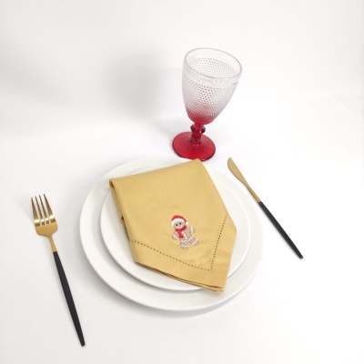 Conjunto de mesa com pratos brancos, guardanapo amarelo bordado, talheres pretos e dourados, copo transparente com pé vermelho.