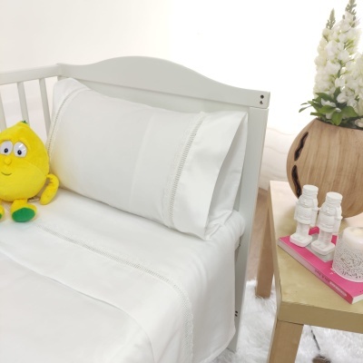 Jogo de cama branco com detalhes bordados num berço branco, peluche amarelo ao lado, mesa com vaso de flores e livros cor-de-rosa